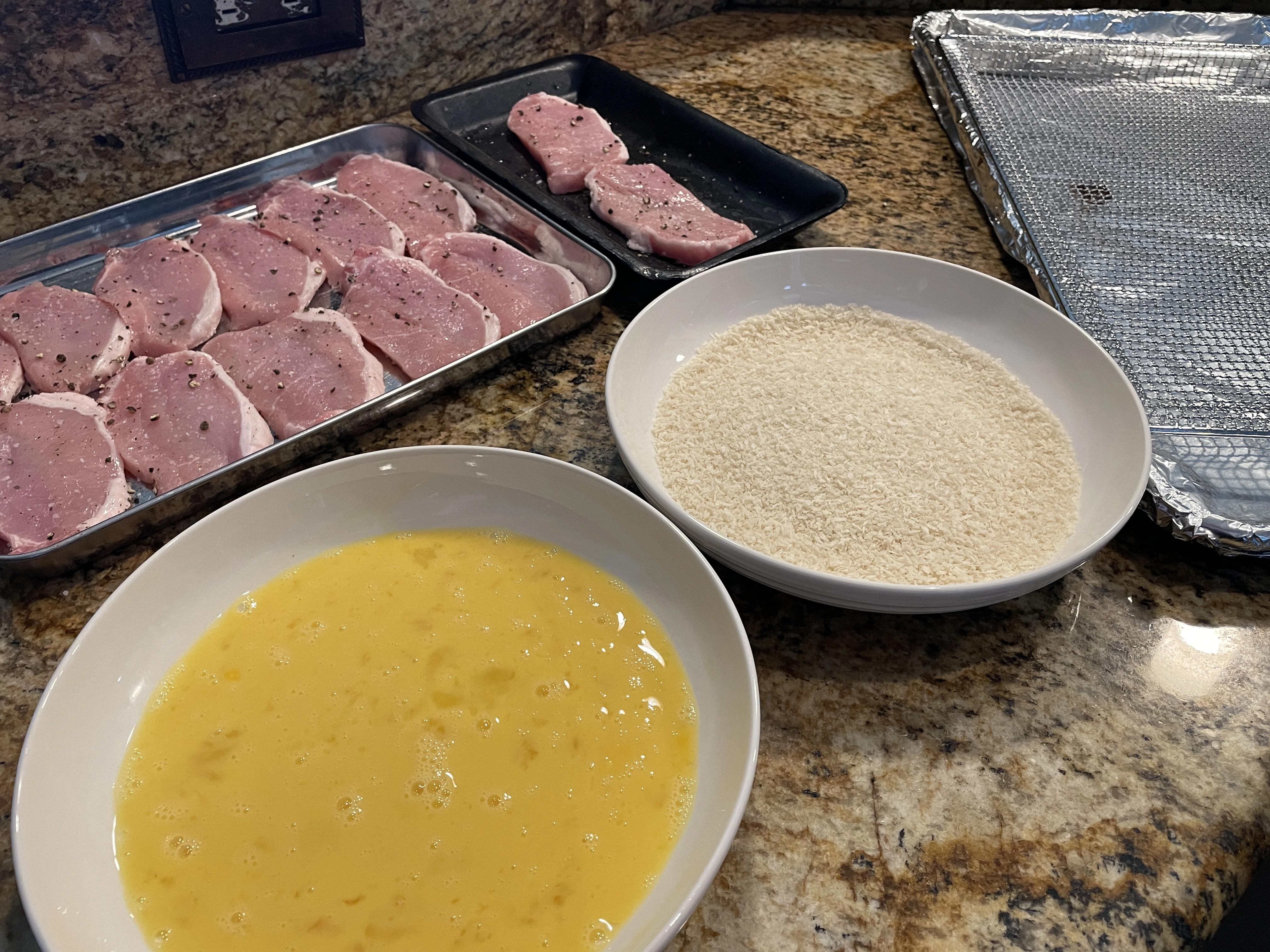 A breading setup on a orange granite countertop. A small sheet pan of raw pork chops seasoned with salt and pepper can be seen in the top left. A wide dish with beaten eggs can be seen in the bottom left, and another wide dish with panko bread crumbs can be seen in the middle. A sheet pan with a wire rack in it is on the right.